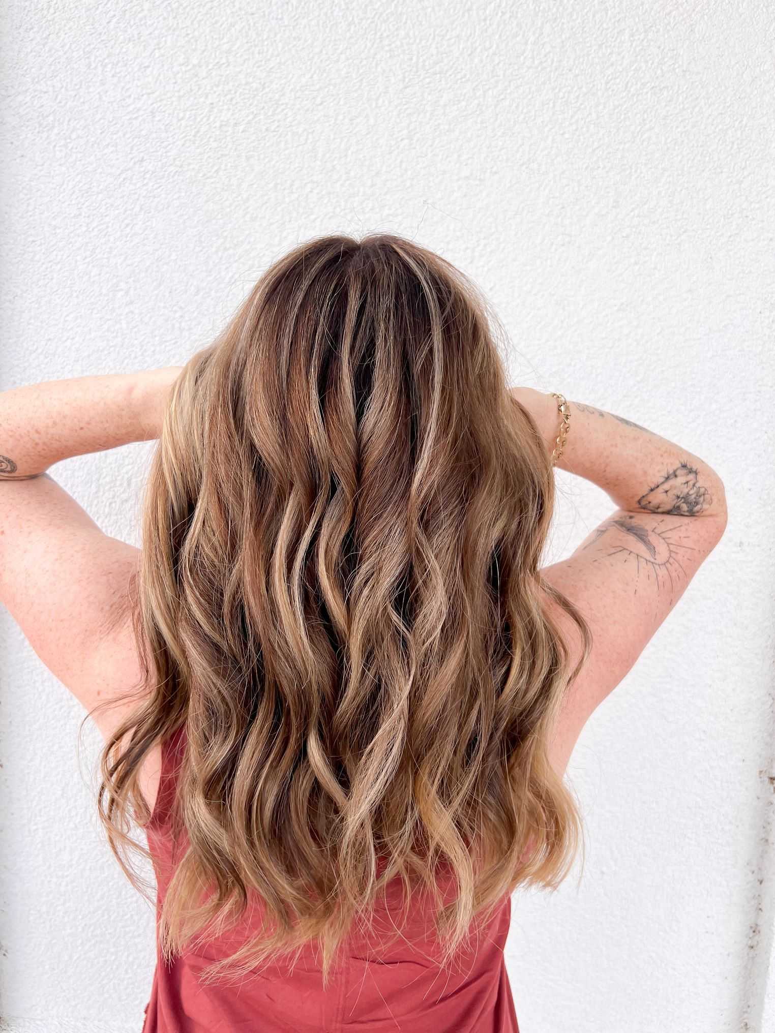 Woman with wavy, highlighted hair, back to the camera, showing tattooed arms in front of a white wall.
