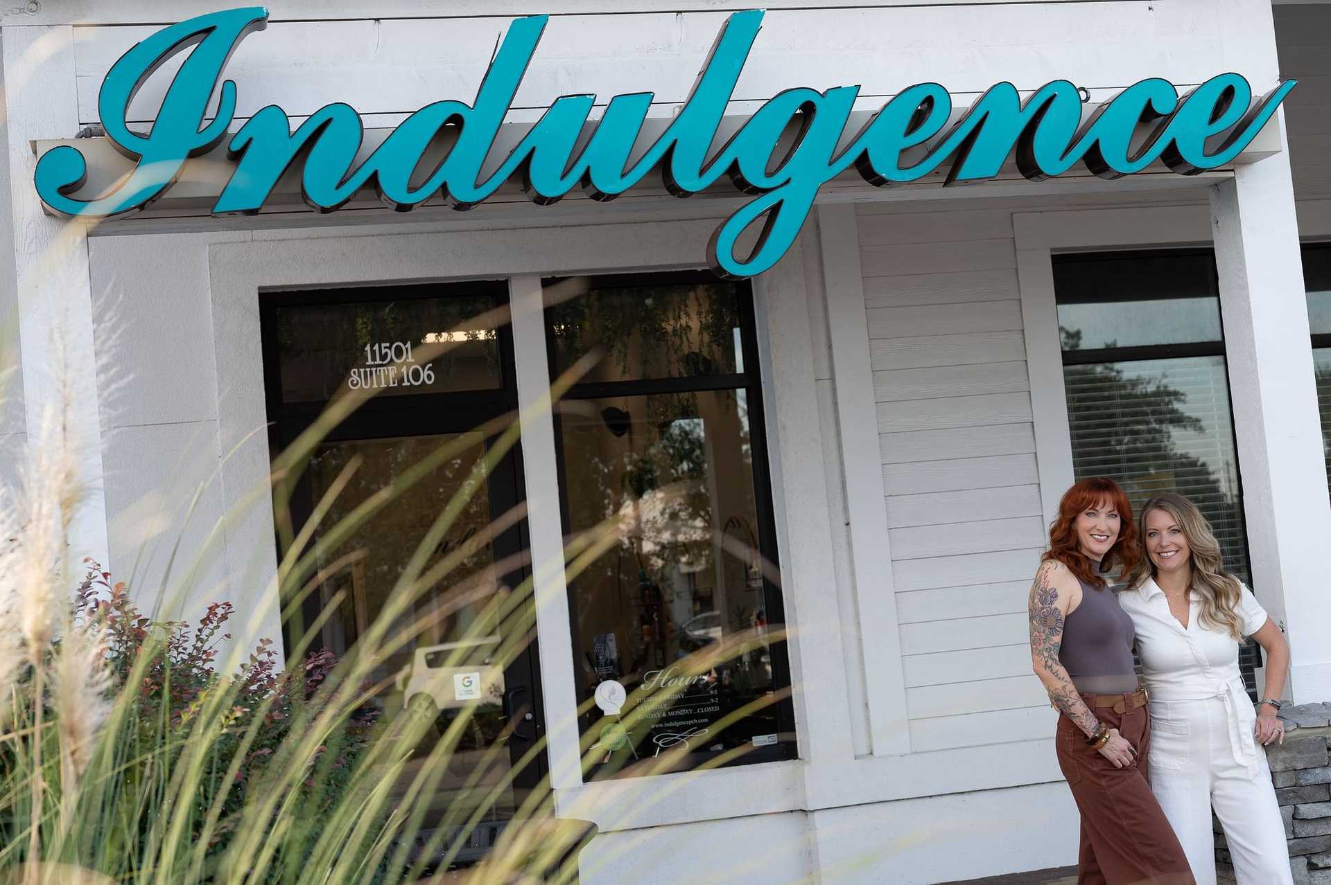 Two women smile and stand in front of a shop named "Indulgence.