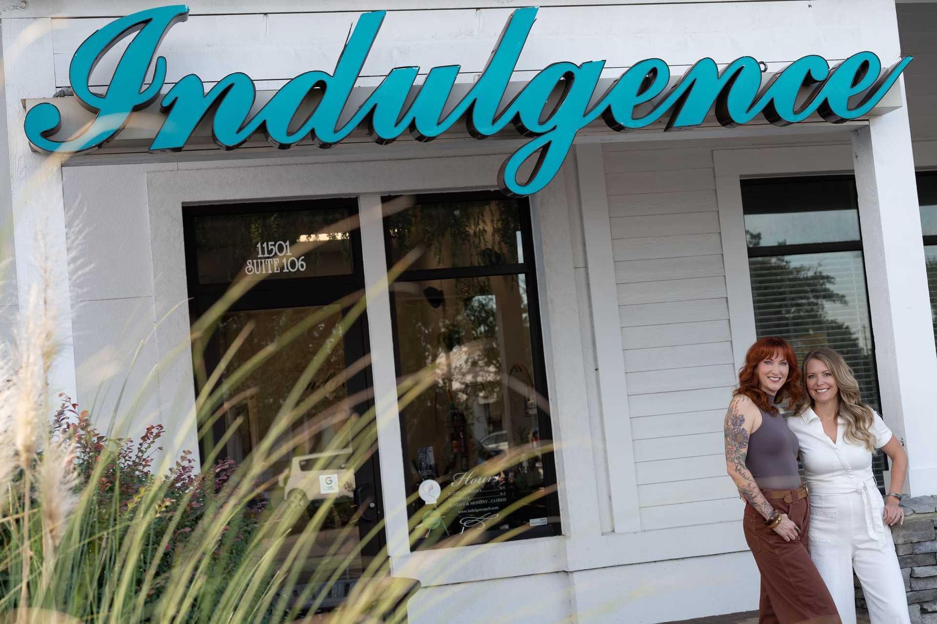 Two women smile and stand in front of a shop named "Indulgence.